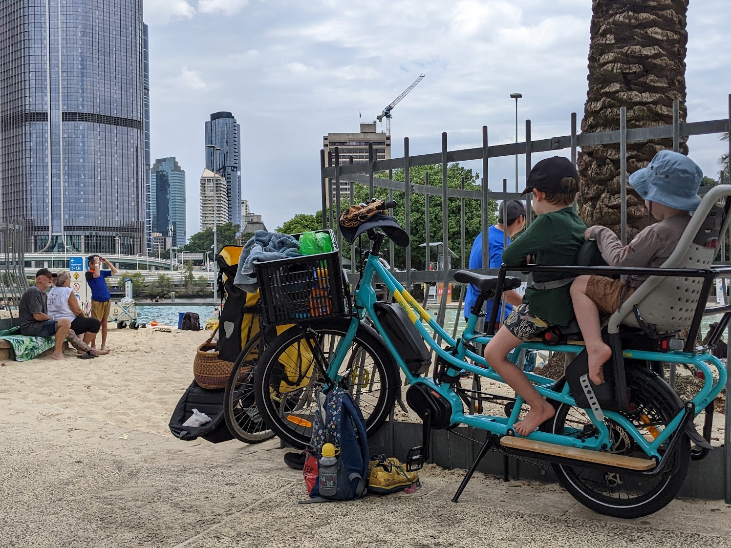 Two children seated on the Tribe Evamos in the foreground facing away from camera. In the background is sand, water, and city buildings (Streets Beach Brisbane).