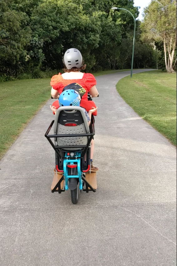 My partner riding, with our two kids aboard. On a bike path in parkland. Rear view.